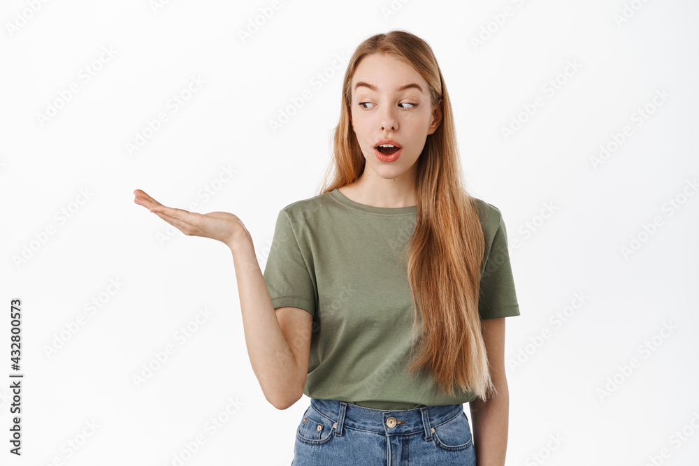 Fototapeta premium Image of excited girl looks at her hand holding copyspace with interest, display item on open palm, standing against white background. Advertisement concept