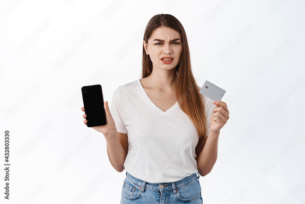 Disappointed frowning female client, showing smartphone and credit card, look frustrated, express dislike, complaining on online shopping app, standing displeased against white background Disappointed frowning female client, showing smartphone and credit card, look frustrated, express dislike, complaining on online shopping app, standing displeased against white background