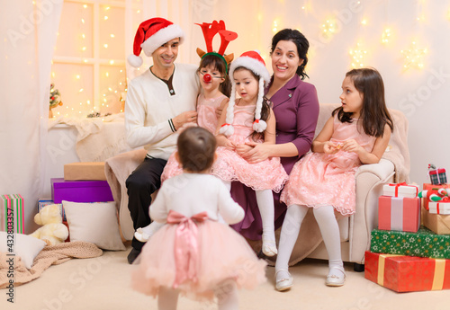 family portrait of a parents and children, dressed in santa helper hat, sitting on a couch in home interior decorated with christmas lights and new year holiday gifts