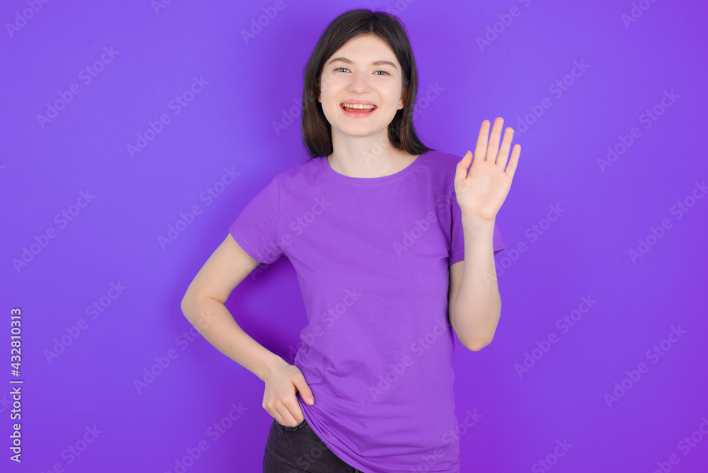 young beautiful Caucasian woman wearing purple T-shirt over purple wall waiving saying hello or goodbye happy and smiling, friendly welcome gesture.