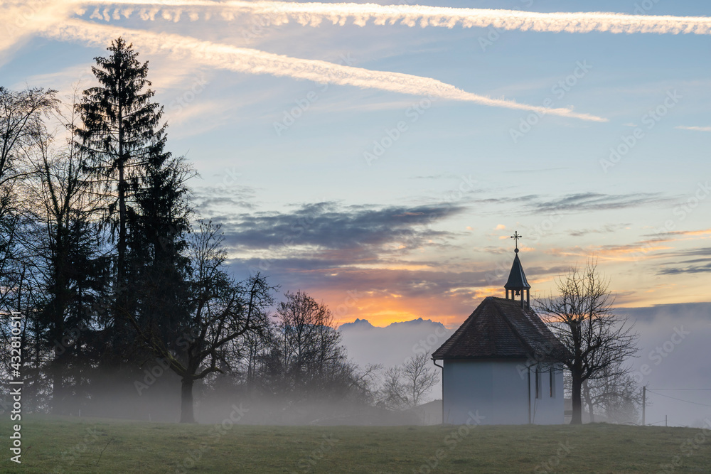Fototapeta premium Kapelle im Winter bei Sonnenuntergang, im Hintergrund das Rheintal mit Nebelmeer und der Säntis im orangen Sonnenlicht. chapel in sunset with sea of fog and colored sky