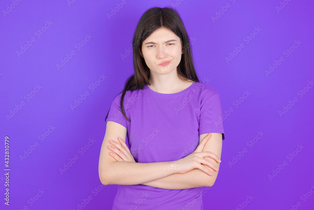 Fototapeta premium Gloomy dissatisfied young beautiful Caucasian girl wearing purple T-shirt over purple background looks with miserable expression at camera from under forehead, makes unhappy grimace