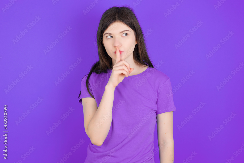 young beautiful Caucasian girl wearing purple T-shirt over purple background makes silence gesture, keeps index finger to lips makes hush sign. Asks not to share secret. young beautiful Caucasian girl wearing purple T-shirt over purple background makes silence gesture, keeps index finger to lips makes hush sign. Asks not to share secret.