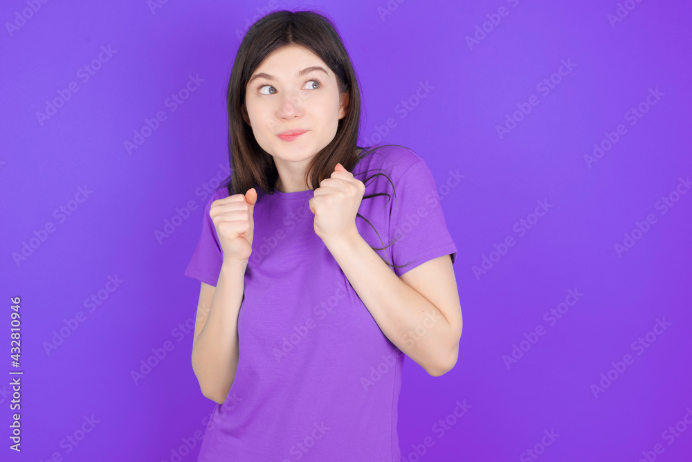 young beautiful Caucasian girl wearing purple T-shirt over purple background clenches fists and awaits for something nice happened looks away bites lips and waits announcement of results young beautiful Caucasian girl wearing purple T-shirt over purple background clenches fists and awaits for something nice happened looks away bites lips and waits announcement of results
