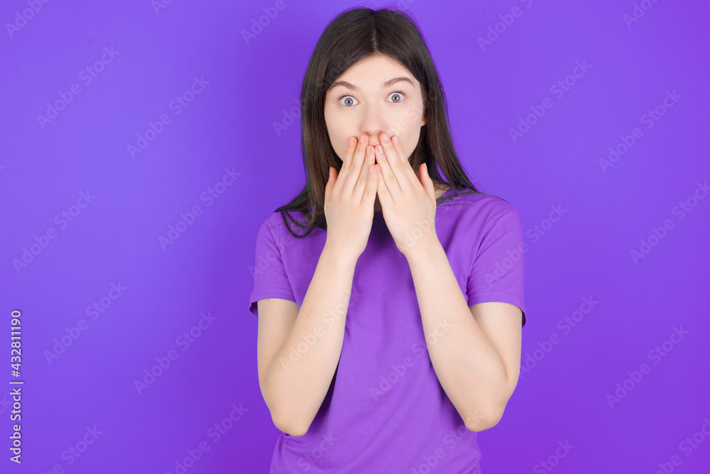 young beautiful Caucasian girl wearing purple T-shirt over purple background keeps hands on mouth, looks with eyes full of disbelief, being puzzled with amount of work young beautiful Caucasian girl wearing purple T-shirt over purple background keeps hands on mouth, looks with eyes full of disbelief, being puzzled with amount of work