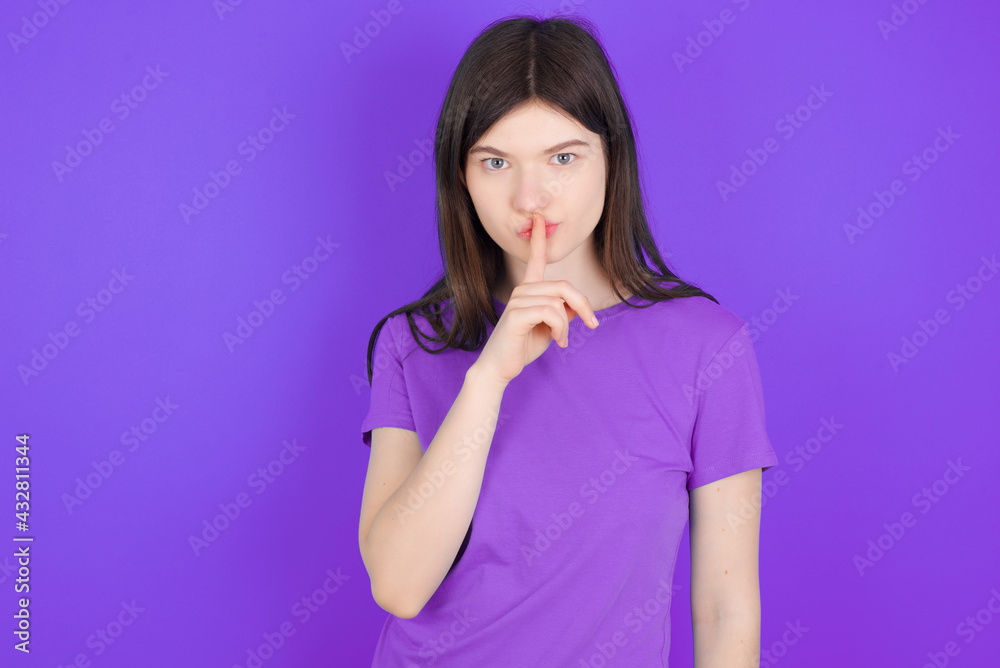 Surprised young beautiful Caucasian girl wearing purple T-shirt over purple background makes silence gesture, keeps finger over lips and looks mysterious at camera Surprised young beautiful Caucasian girl wearing purple T-shirt over purple background makes silence gesture, keeps finger over lips and looks mysterious at camera