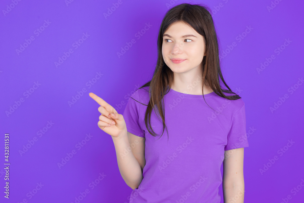 young beautiful Caucasian girl wearing purple T-shirt over purple background points at copy space and advertises something, advices best price. young beautiful Caucasian girl wearing purple T-shirt over purple background points at copy space and advertises something, advices best price.