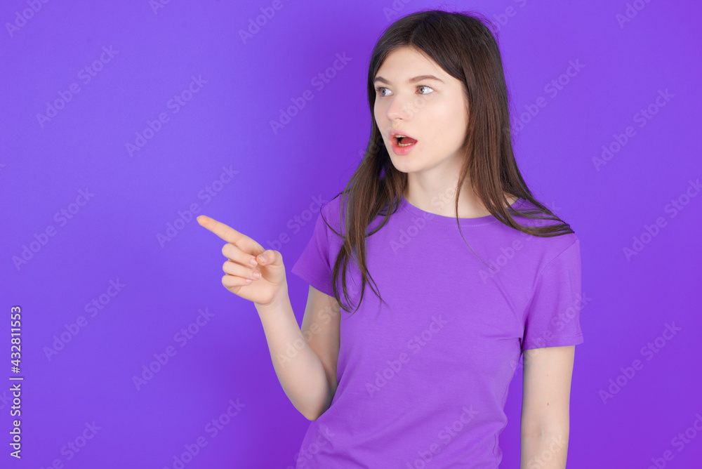 Stunned young beautiful Caucasian girl wearing purple T-shirt over purple background with greatly surprised expression points away on copy space, indicates something Stunned young beautiful Caucasian girl wearing purple T-shirt over purple background with greatly surprised expression points away on copy space, indicates something