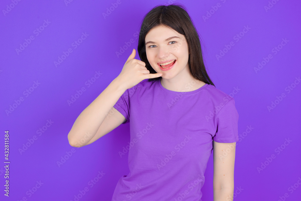 young beautiful Caucasian girl wearing purple T-shirt over purple background makes phone gesture, says call me back again, has glad expression. young beautiful Caucasian girl wearing purple T-shirt over purple background makes phone gesture, says call me back again, has glad expression.
