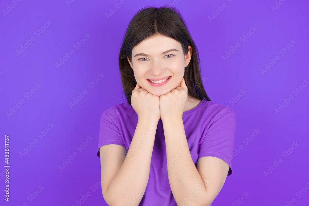 Satisfied young beautiful Caucasian girl wearing purple T-shirt over purple background touches chin with both hands, smiles pleasantly, rejoices good day with lover Satisfied young beautiful Caucasian girl wearing purple T-shirt over purple background touches chin with both hands, smiles pleasantly, rejoices good day with lover