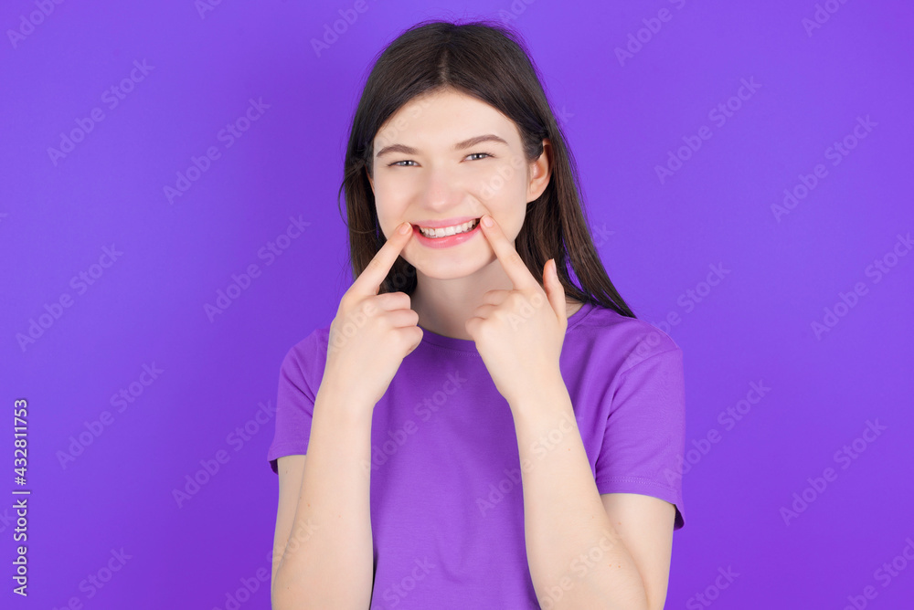 Happy young beautiful Caucasian girl wearing purple T-shirt over purple background with toothy smile, keeps index fingers near mouth, fingers pointing and forcing cheerful smile Happy young beautiful Caucasian girl wearing purple T-shirt over purple background with toothy smile, keeps index fingers near mouth, fingers pointing and forcing cheerful smile