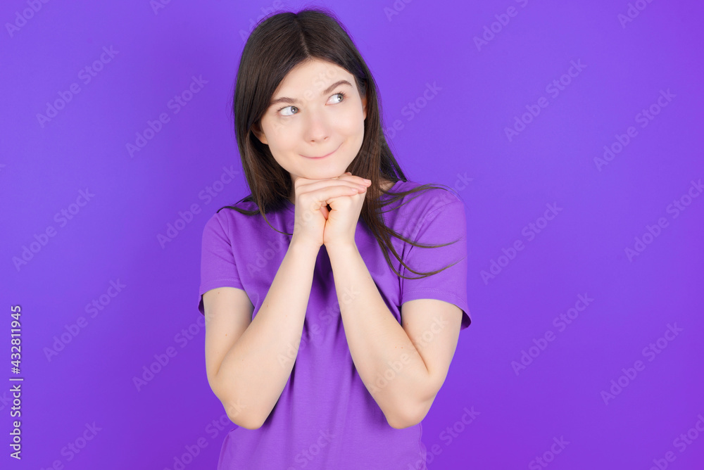 Fototapeta premium Curious young beautiful Caucasian girl wearing purple T-shirt over purple background keeps hands under chin bites lips and looks with interest aside.