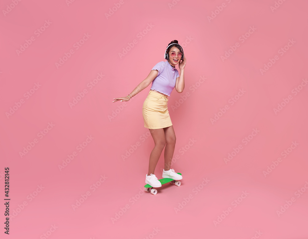 Young asian teenage girl playing on skateboard with wearing wireless headphones listening to music on pink background. Young asian teenage girl playing on skateboard with wearing wireless headphones listening to music on pink background.