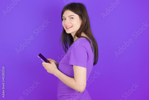 Rear view photo portrait of young beautiful Caucasian girl wearing purple T-shirt over purple background using smartphone smiling