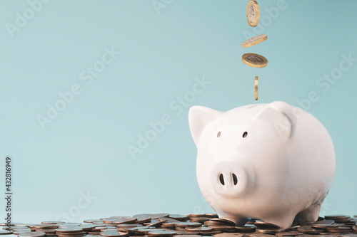 Slika na platnu Coins falling and dropping to white piggy bank saving on top of heap of coins and blue background ,money saving for investment and retirement in the future concept