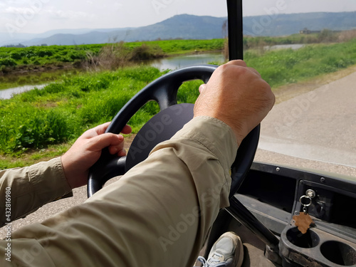 Driving golf car on nature. Man steers electric car. Driver hands are on a steering wheel. Green meadow and water canal view along the road. Mountains on skyline 