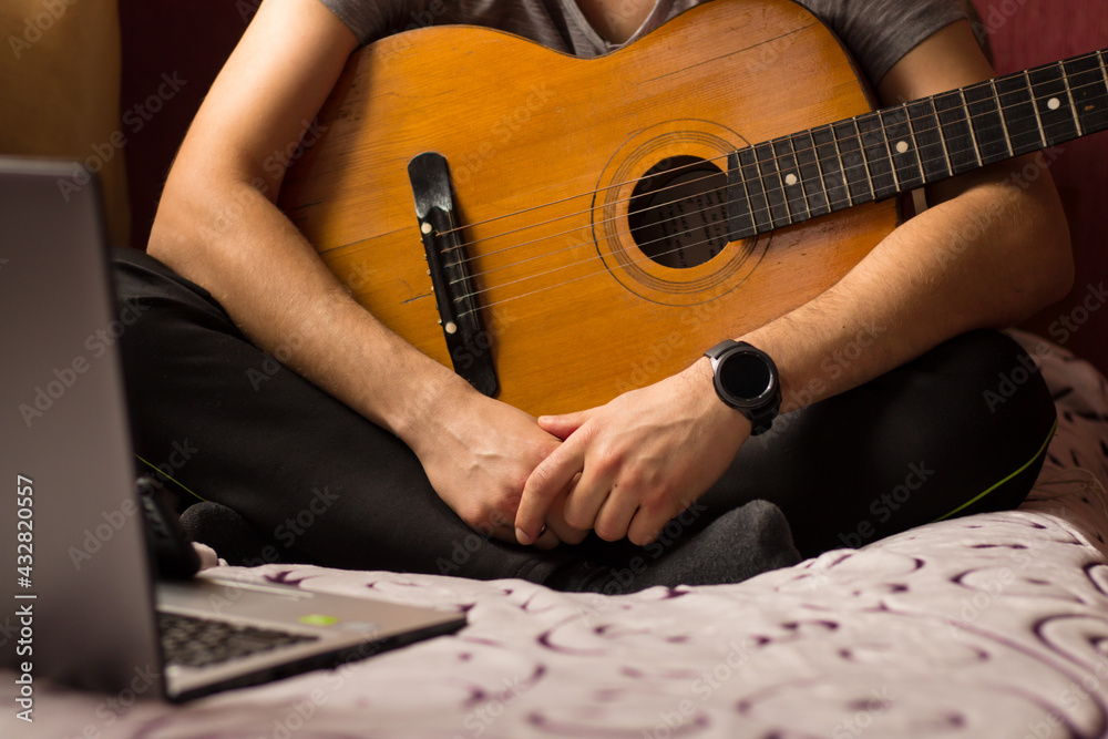 Fototapeta premium Learn to play the guitar online at home. A young guy with a guitar sits on the bed in front of a laptop and watches online guitar learning.