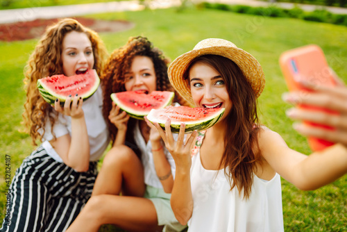 Three young woman relaxing on the grass, eating watermelon and taking selfie on the phone. People, lifestyle, travel, nature and vacations concept.
