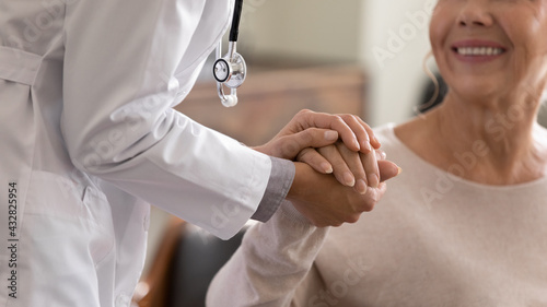 Doctor giving hope. Close up shot of young female physician leaning forward to smiling elderly lady patient holding her hand in palms. Woman caretaker in white coat supporting encouraging old person photo