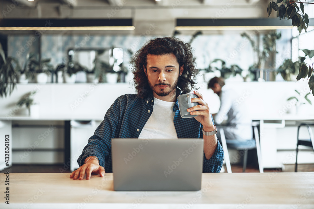 Young businessman is using laptop and holding a cup of coffee in office - obrazy, fototapety, plakaty 