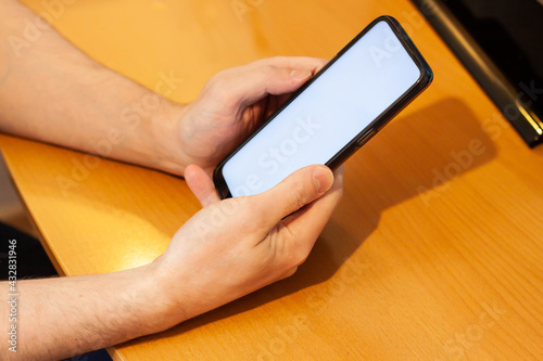 Blank white screen cell phone. Male hand texting using a mobile phone on a wooden table. Background empty space for advertising text.