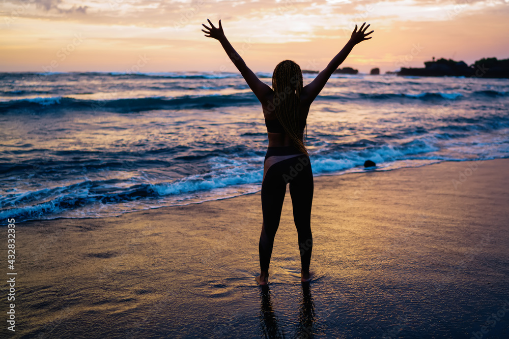 Back view of satisfied female runner in sportive tracksuit raising hands during workout break at seashore coastline, carefree woman with casual figure enjoying slimming goals and own triumph