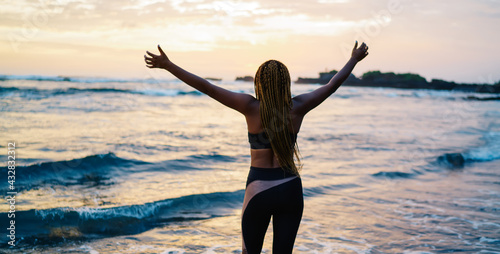 Back view of satisfied female runner in sportive tracksuit raising hands during workout break at seashore coastline  carefree woman with casual figure enjoying slimming goals and own triumph