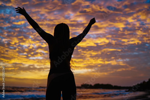 Back silhouette view of satisfied fit girl celebrating sportive goals during evening time at coastline with picturesque colorful sky, carefree female athlete with casual figure feeling freedom