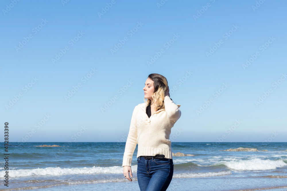 young woman walking along the beach on a sunny day, wearing jeans and a wool sweater