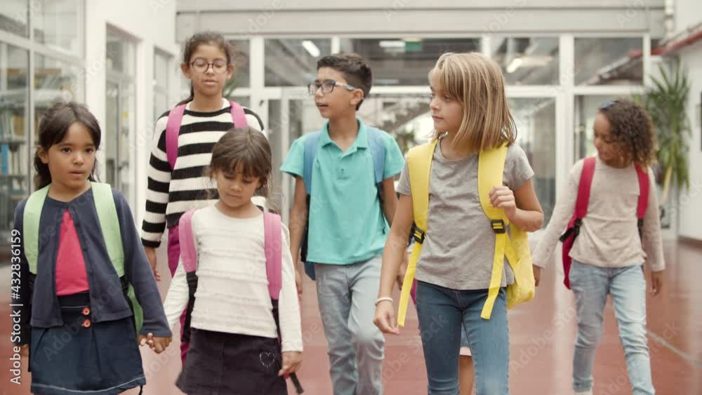 Friendly children with colorful backpacks walking in school hallway. Boy and girl in eyeglasses talking to each other. Little girls holding hands. Back to school concept. Friendly children with colorful backpacks walking in school hallway. Boy and girl in eyeglasses talking to each other. Little girls holding hands. Back to school concept.