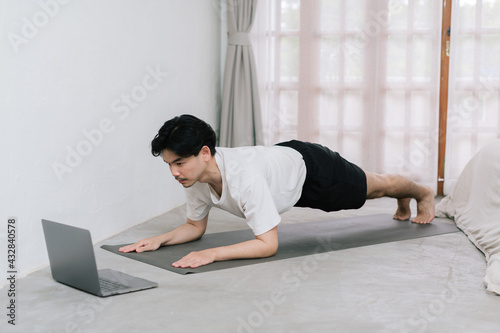 Training At Home. Sporty man doing yoga while watching online tutorial on laptop, exercising in living room, free space