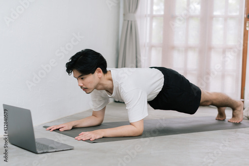 Training At Home. Sporty man doing yoga while watching online tutorial on laptop, exercising in living room, free space