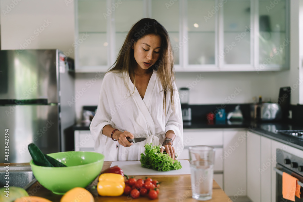 Stylish young ethnic woman cutting veggies while cooking in light kitchen