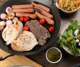 Mix meet chicken beef steak sausage tomato onion garlic fry grill oil metal fry pan colourful pasta green salad with cheese olive mint basil pesto sauce board knife fork spoon rustic wooden table