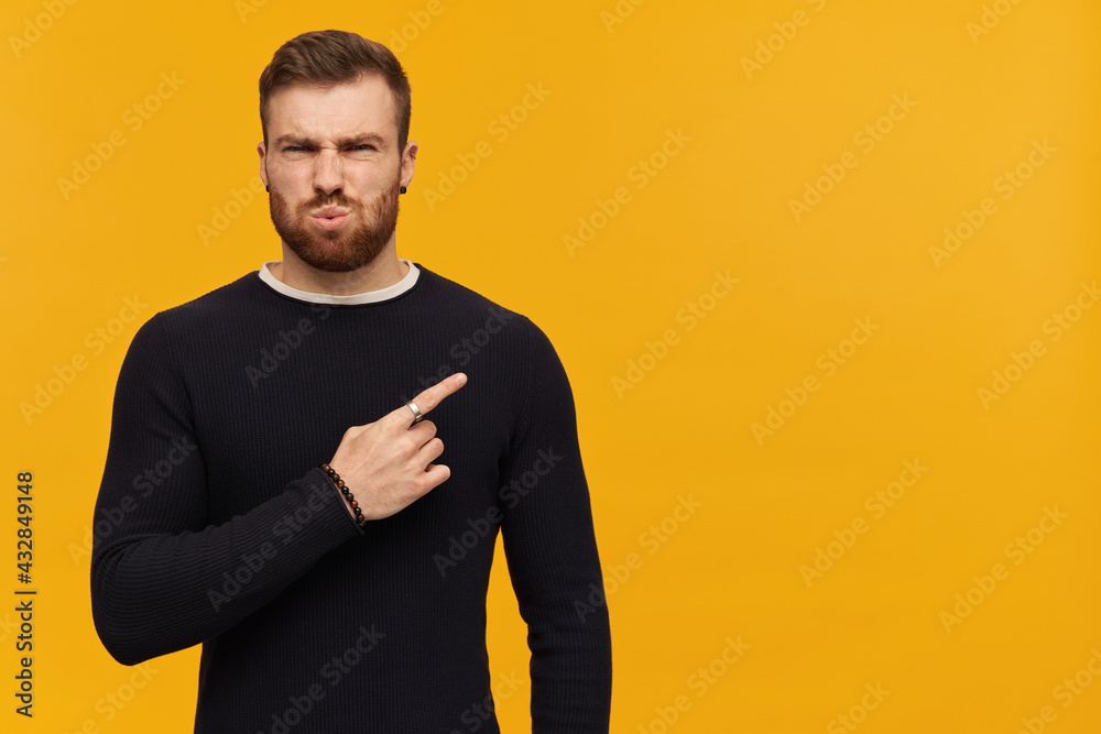Portrait of annoyed, squinting male with brunette hair and beard. Wearing black sweater. Watching at the camera and pointing finger to the right at copy space, isolated over yellow background Portrait of annoyed, squinting male with brunette hair and beard. Wearing black sweater. Watching at the camera and pointing finger to the right at copy space, isolated over yellow background