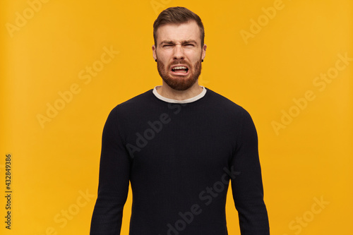 Portrait of upset, crying male with brunette hair and bristle. Has piercing. Wearing black sweater. Emotion concept. Watching at the camera, isolated over yellow background