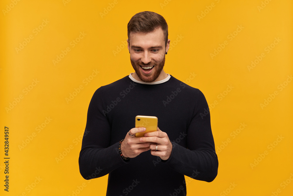Excited looking male, handsome guy with brunette hair and beard. Has piercing. Wearing black sweater. Holds smartphone and watching at it. Reading a message. Stand isolated over yellow background Excited looking male, handsome guy with brunette hair and beard. Has piercing. Wearing black sweater. Holds smartphone and watching at it. Reading a message. Stand isolated over yellow background