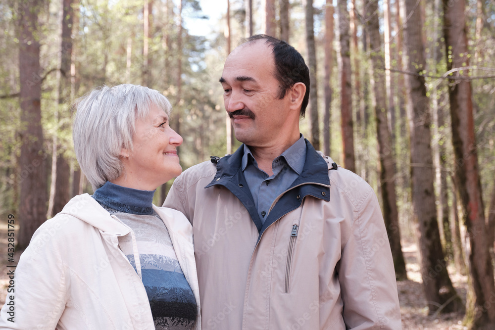 Elderly interracial couple in a spring forest park, smiling, looking at each other.