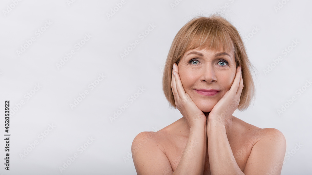 Lovely senior woman with soft skin looking at camera, touching face on light background, banner design with copy space Lovely senior woman with soft skin looking at camera, touching face on light background, banner design with copy space