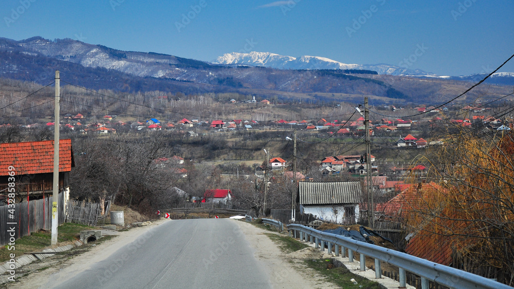 Buila mountain crest rises above a rural settlement with houses scattered along the neighboring hills. Springtime, snow covers the mountain peaks. Romania, Carpathia.