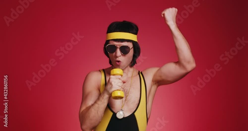 Funny retro athlete man in yellow tracksuit and sunglasses from 80's with dumbbells sings like a rock star isolated on red background. Retro sport hipster show off and posing in front of camera. photo