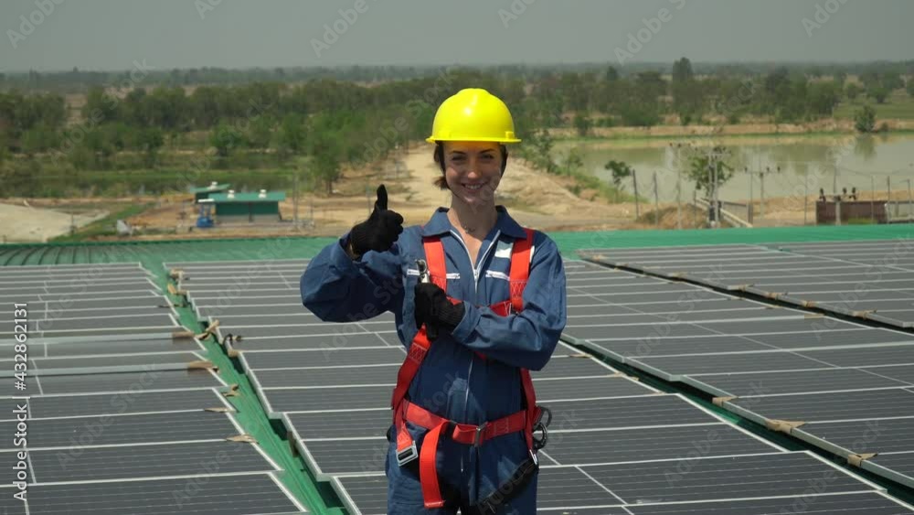 Solar power plant engineer woman in hard hat show Thumbs up smiling with photovoltaic on roof top at warehouse factory. technician female worker standing on Solar panels and looking camera outdoors.