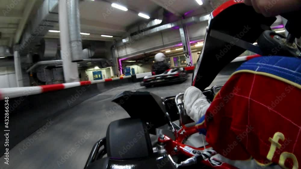 Two kart driver wearing helmets and racing suits drive over an indoor track and quickly around tight curves - on board camera on second cart