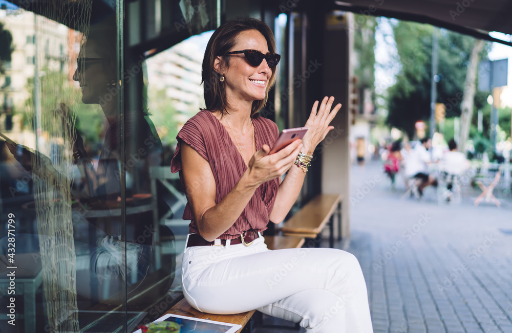 Fototapeta premium Beautiful female blogger surprised with success of publication gesture emotionally holding mobile phone, smiling caucasian woman in trendy clothes and sunglasses laughing feeling confused from message