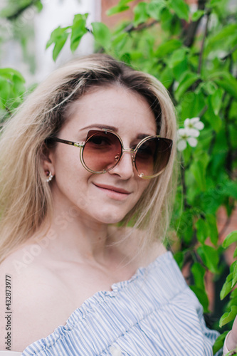 Young blonde in sunglasses poses in the foliage of a blossoming tree