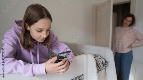 Fotografie Addicted to modern technology young teen girl sitting on bed involved in communicating in social network with friends using smartphone, dissatisfied worried mother on background, parental control