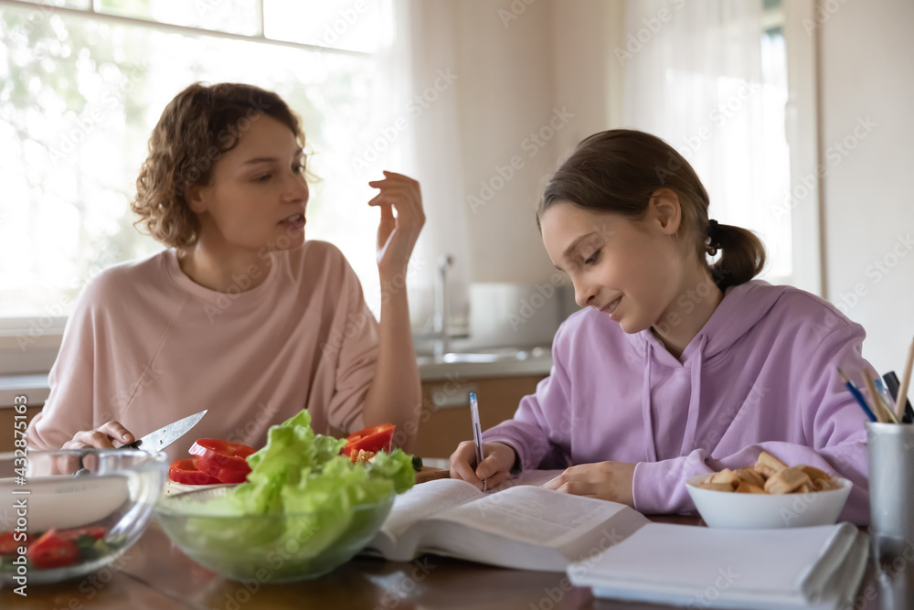 Happy mother helping young teen child daughter with learning, preparing assignment, school task or homework while cooking healthy vegetarian salad for dinner in kitchen, casual daily routine concept. Happy mother helping young teen child daughter with learning, preparing assignment, school task or homework while cooking healthy vegetarian salad for dinner in kitchen, casual daily routine concept.