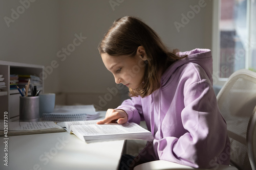 Concentrated young teenage girl in comfort hoodie reading educational material from textbook, learning foreign language or preparing alone for school examination, study at home, homeschooling concept.