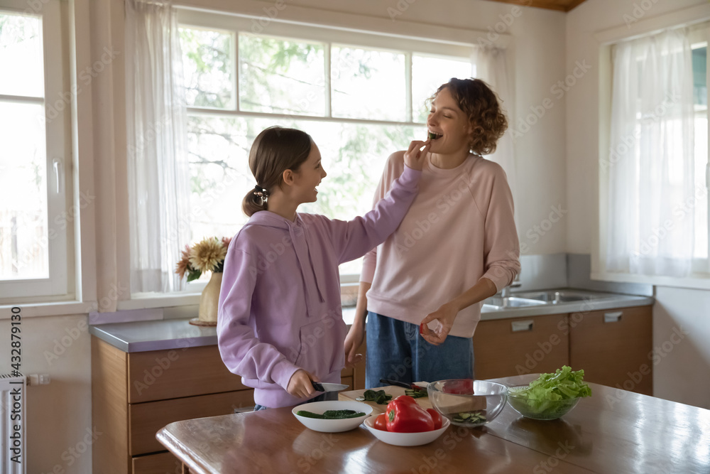 Smiling young teen kid girl feeding happy mother, preparing healthy food together in modern kitchen. Joyful sincere laughing different generations family enjoying cooking on weekend, dietary concept.