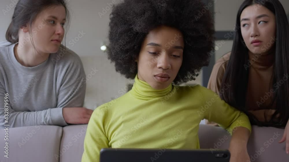 Portrait of absorbed African American young woman messaging online on tablet with bored Caucasian and Asian friends at background. Concentrated intelligent lady ignoring guests working in home office Portrait of absorbed African American young woman messaging online on tablet with bored Caucasian and Asian friends at background. Concentrated intelligent lady ignoring guests working in home office